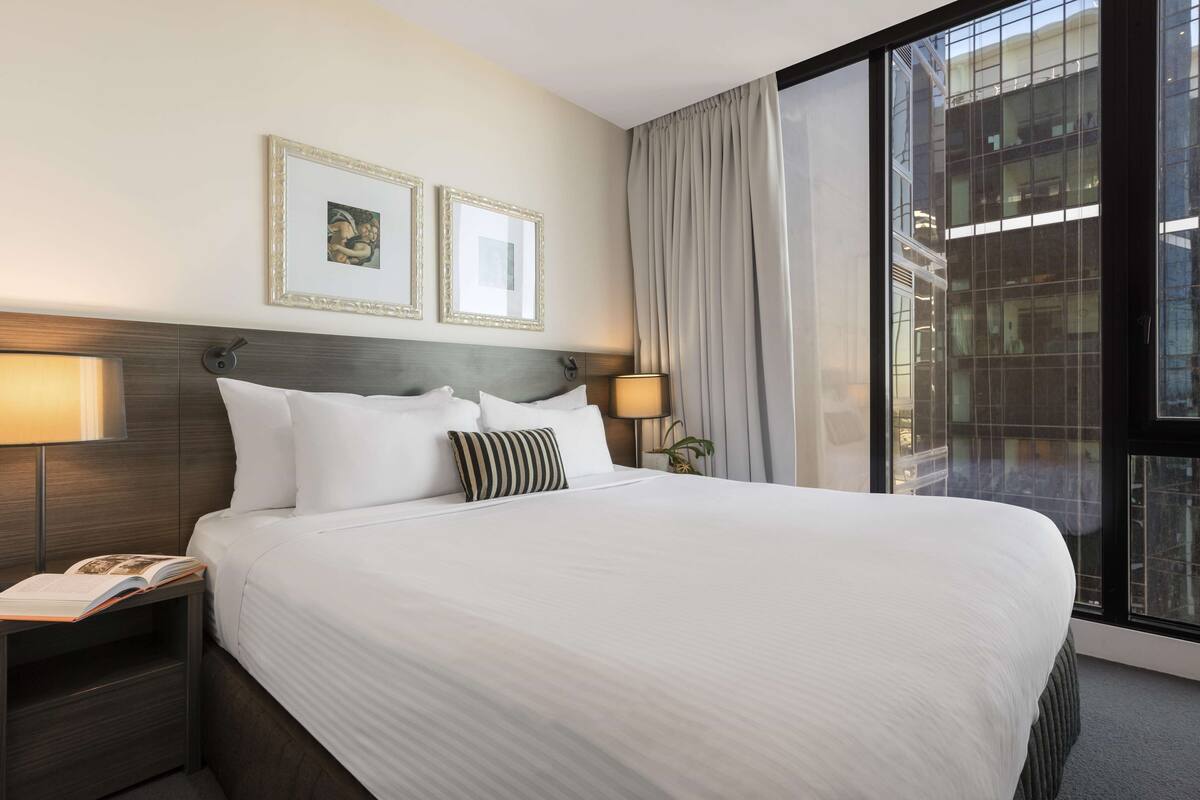 A hotel room with a large bed, two framed pictures on the wall, a bedside lamp, and a window with curtains.
