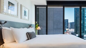 A modern hotel room with a large bed, two framed pictures, a nightstand with a lamp, and a view of the cityscape. - WRAP on Southbank (Southbank)