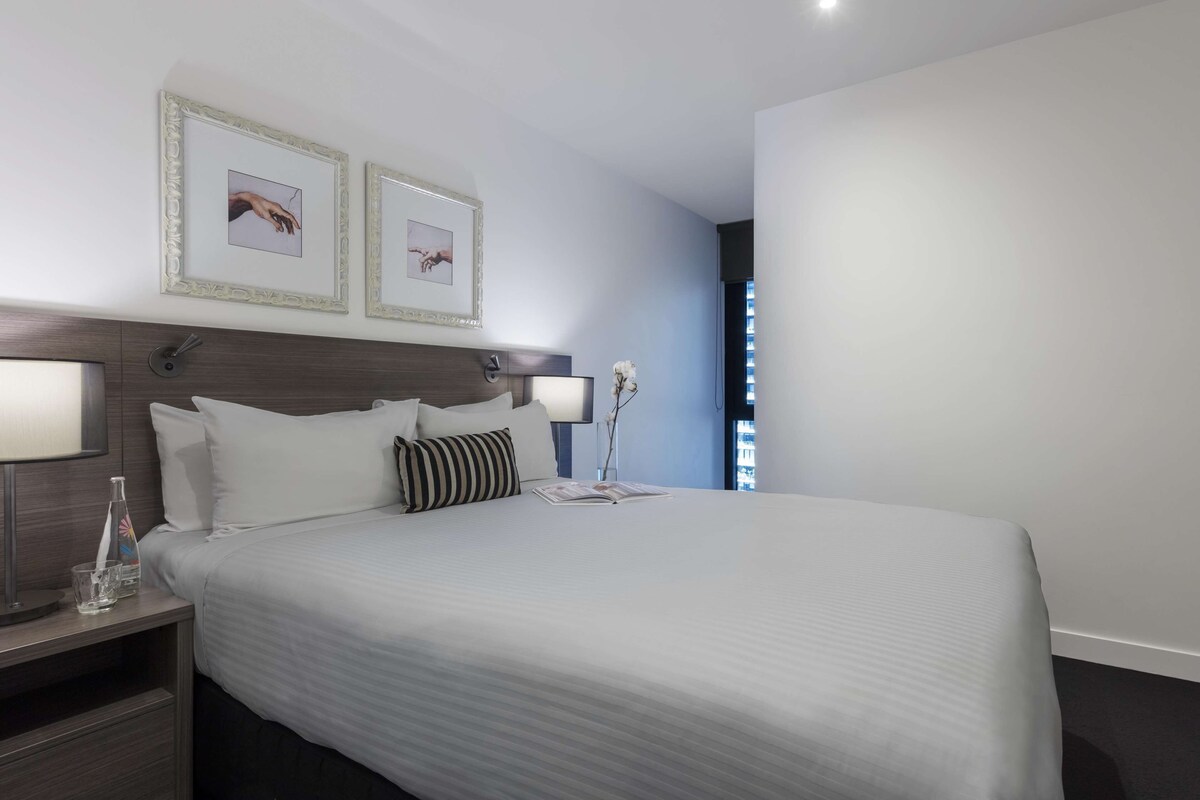 A neatly made bed with white linens, two framed pictures on the wall, a bedside lamp, and a vase with a single flower.