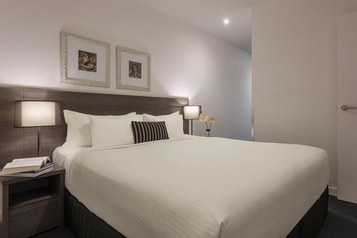 A hotel room with a large bed, two bedside lamps, a nightstand with a book, and two framed pictures on the wall.