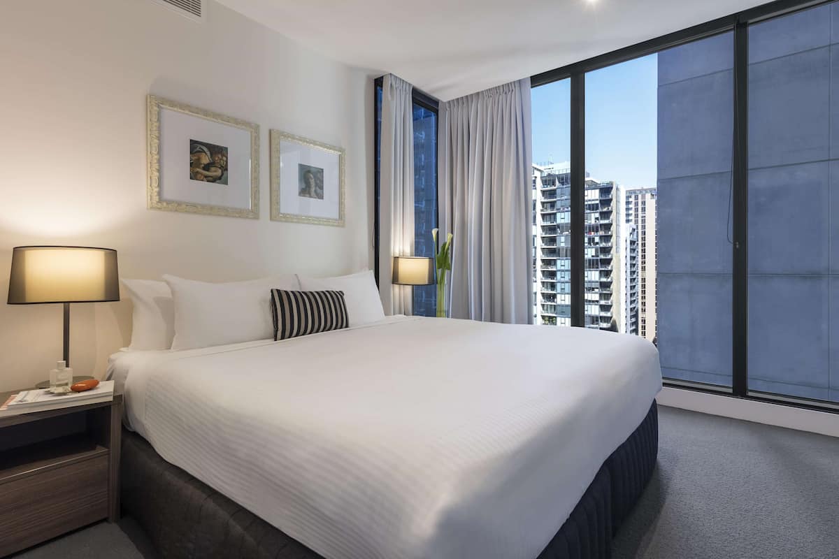 A hotel room with a large bed, two bedside tables with lamps, a window with curtains, and framed pictures on the wall.