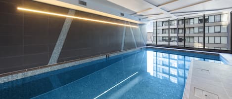A modern indoor swimming pool with clear blue water, a long, narrow shape, and a ceiling with recessed lighting.
