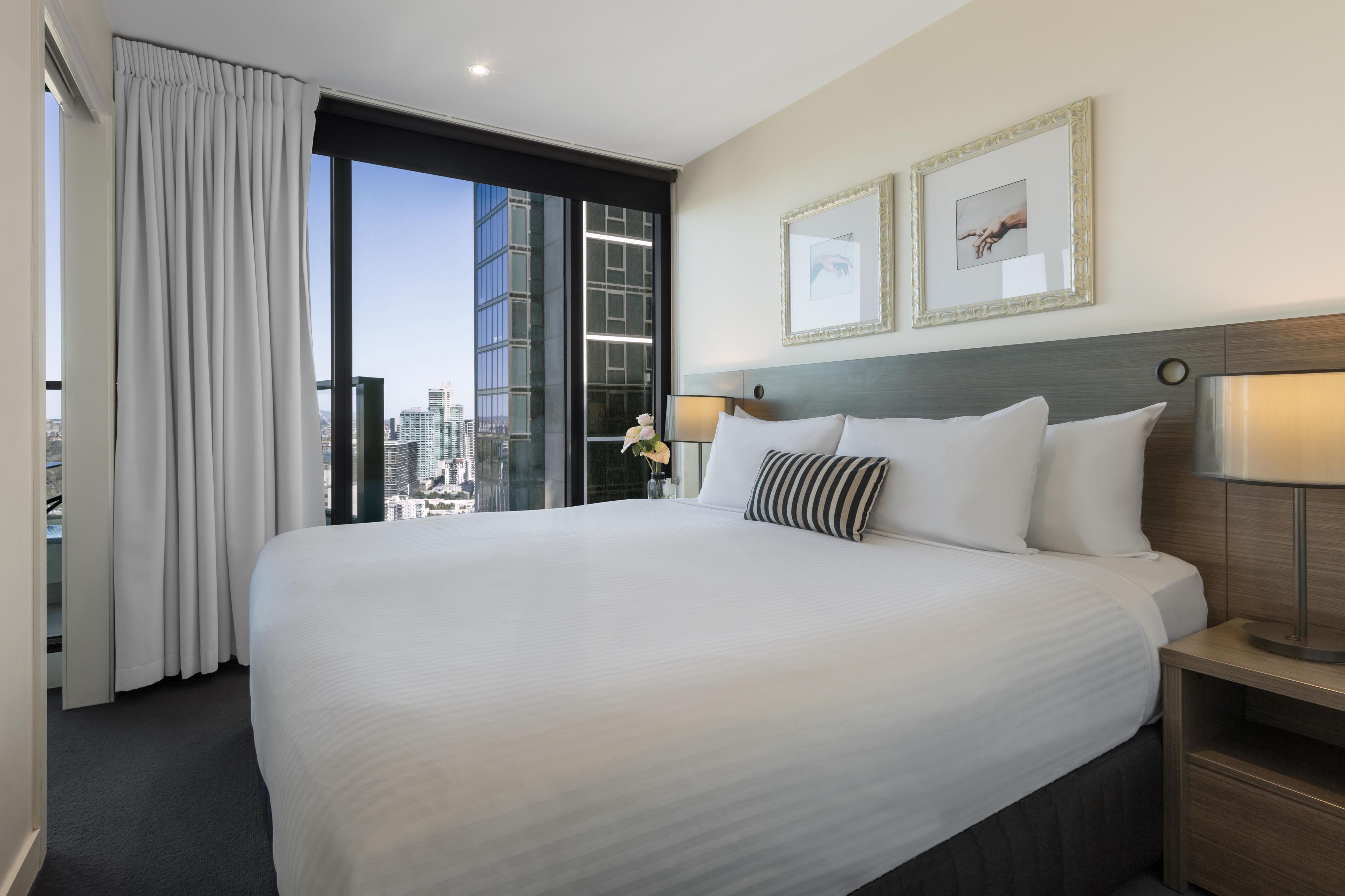 A hotel room with a large bed, two bedside lamps, a nightstand, a vase with flowers, and a city view through floor-to-ceiling windows.
