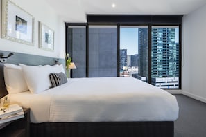A modern hotel room with a large bed, a nightstand with a lamp, framed pictures on the wall, and a view of a cityscape through floor-to-ceiling windows. - WRAP on Southbank (Southbank)