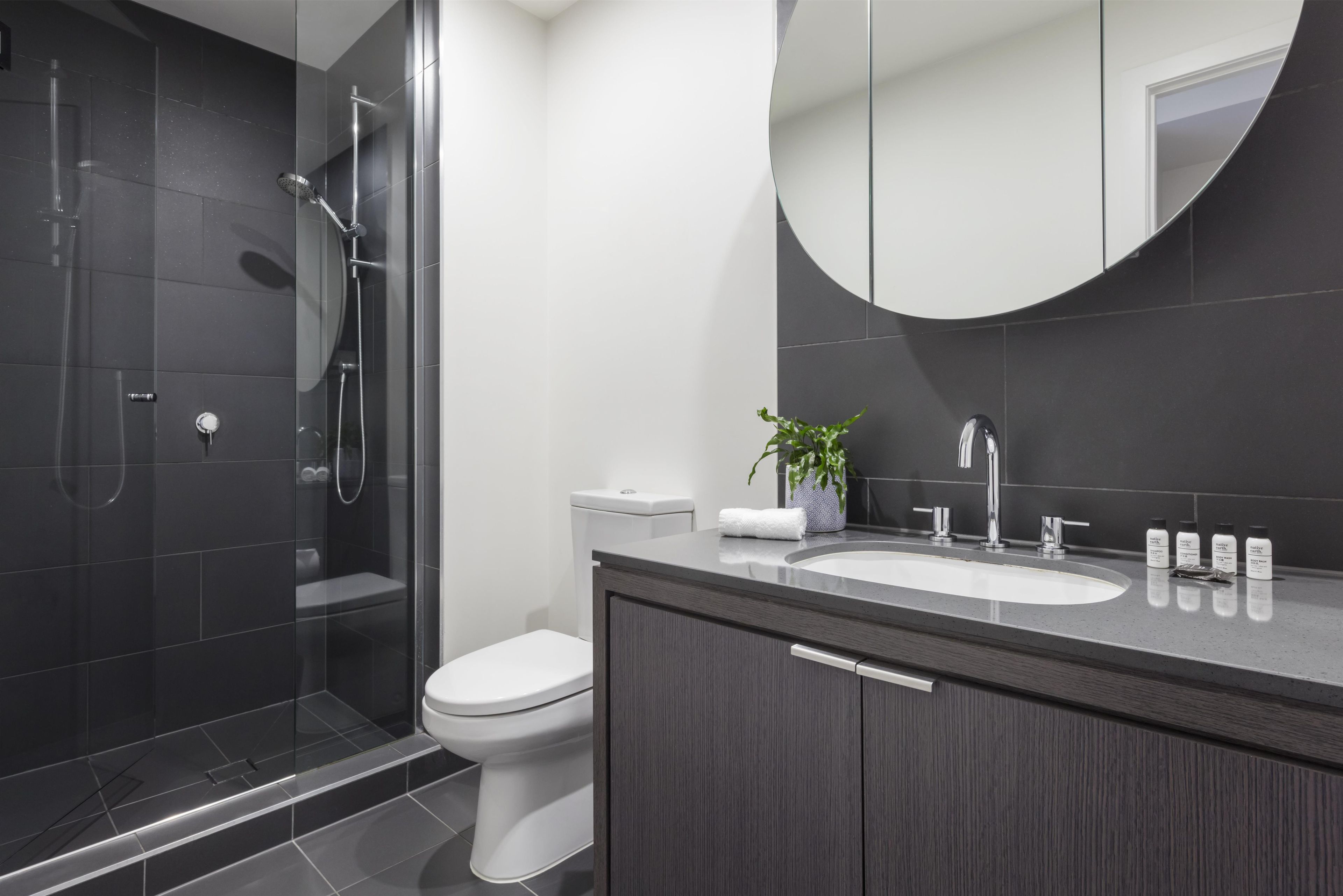 A modern bathroom with a glass-enclosed shower, a white toilet, a dark wood vanity, and a large mirror.
