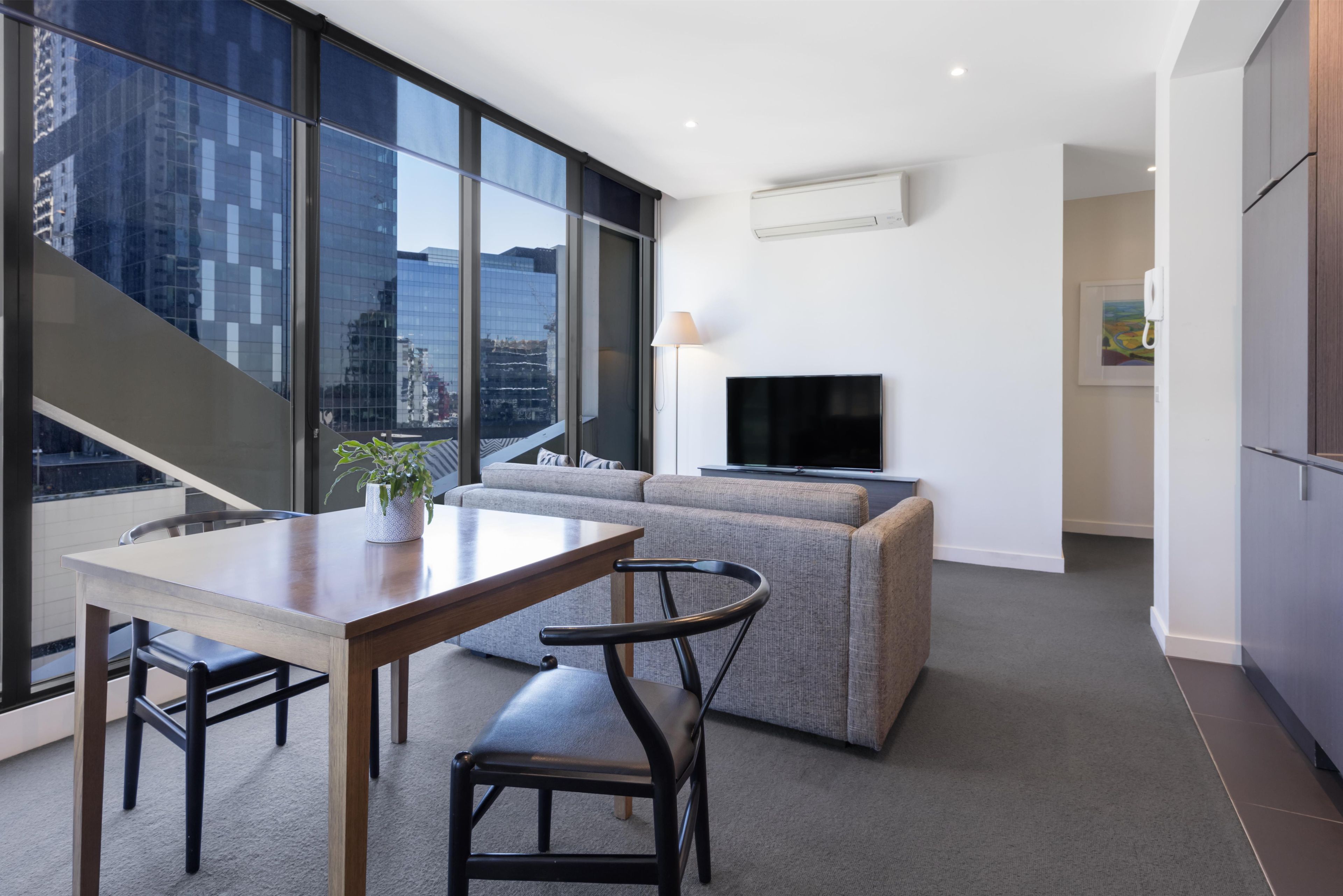 A modern living room with a sofa, a dining table, and chairs, a flat-screen TV mounted on the wall, and a large window offering a city view.