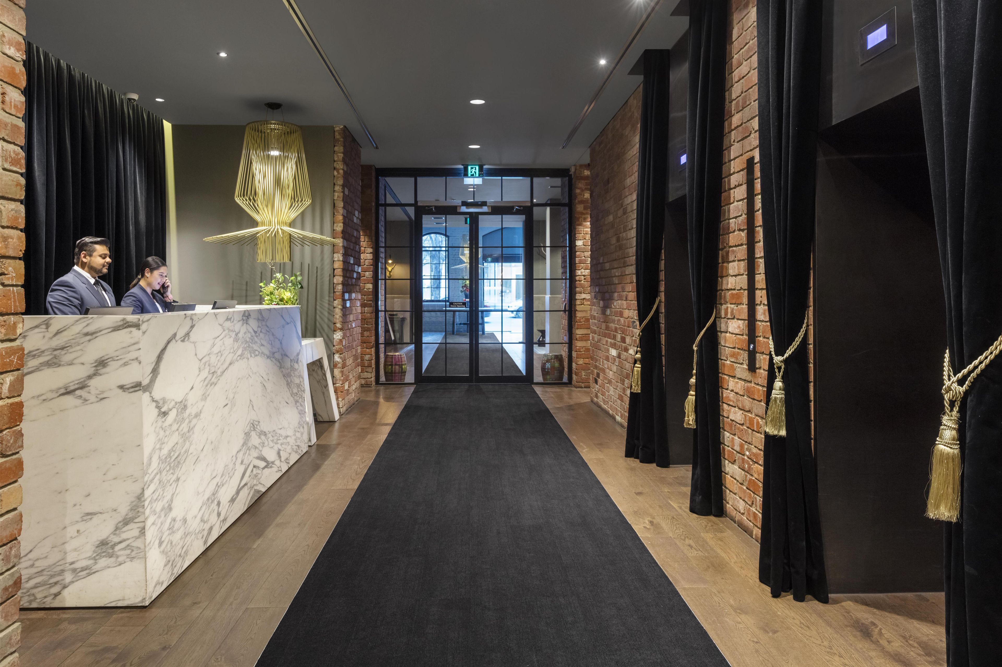 Un área de recepción con mostrador de mármol, pasillo con alfombra negra, paredes de ladrillo y una lámpara moderna.