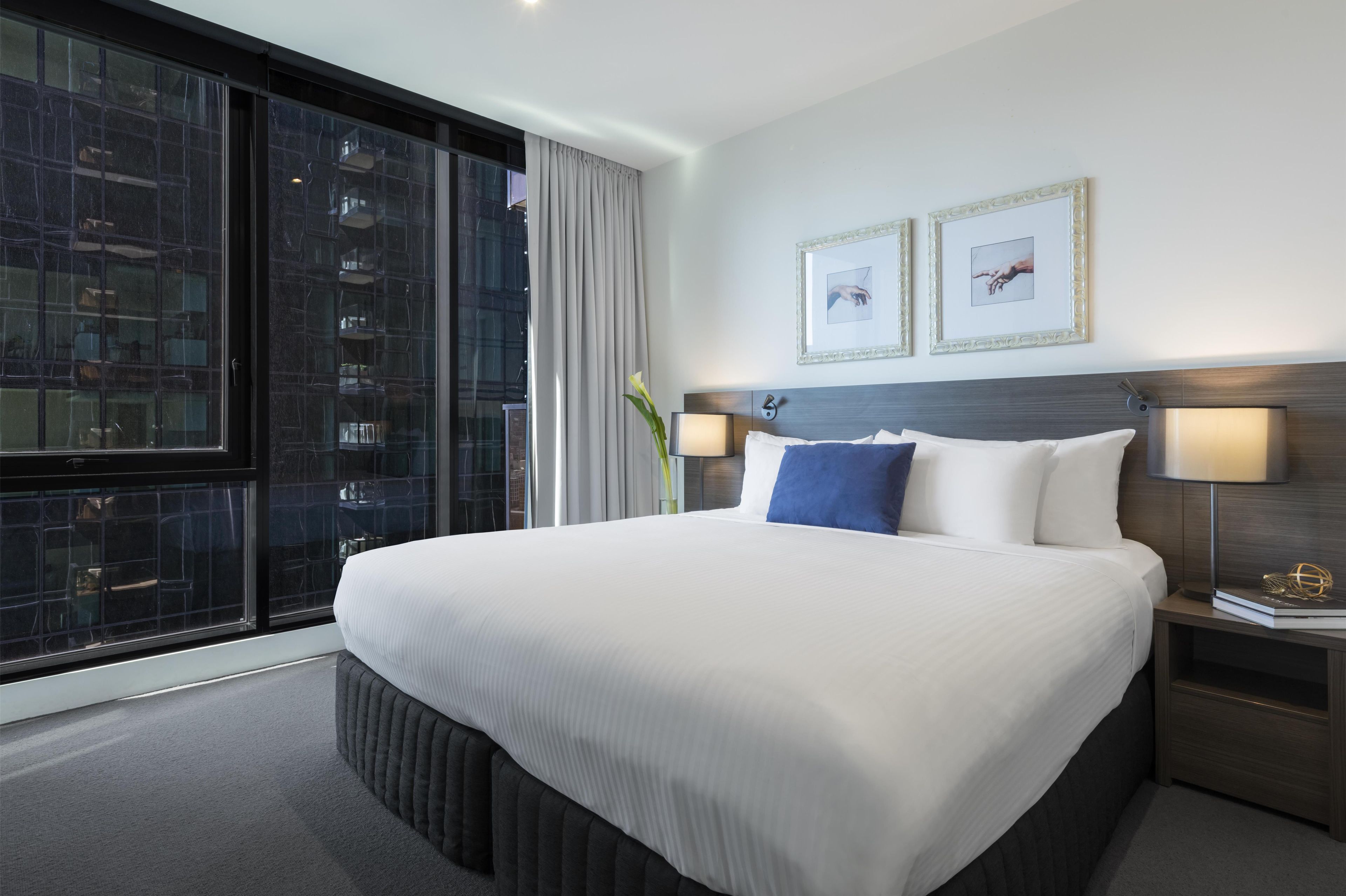 A hotel room with a large bed, two bedside tables with lamps, a window with curtains, and two framed pictures on the wall.