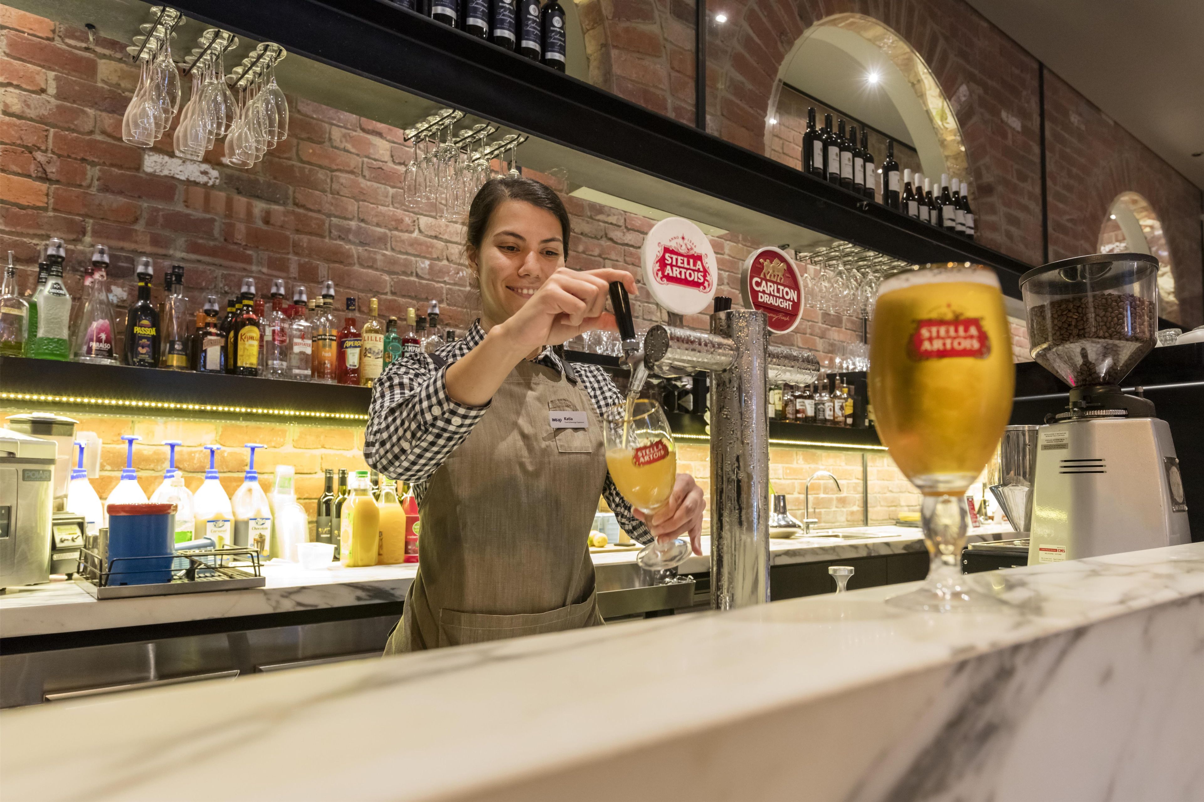 A bartender pouring a drink into a glass at a bar with a brick wall and shelves of liquor bottles.
