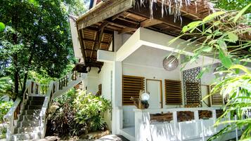 A white building with a wooden staircase leading to a balcony, surrounded by lush greenery.