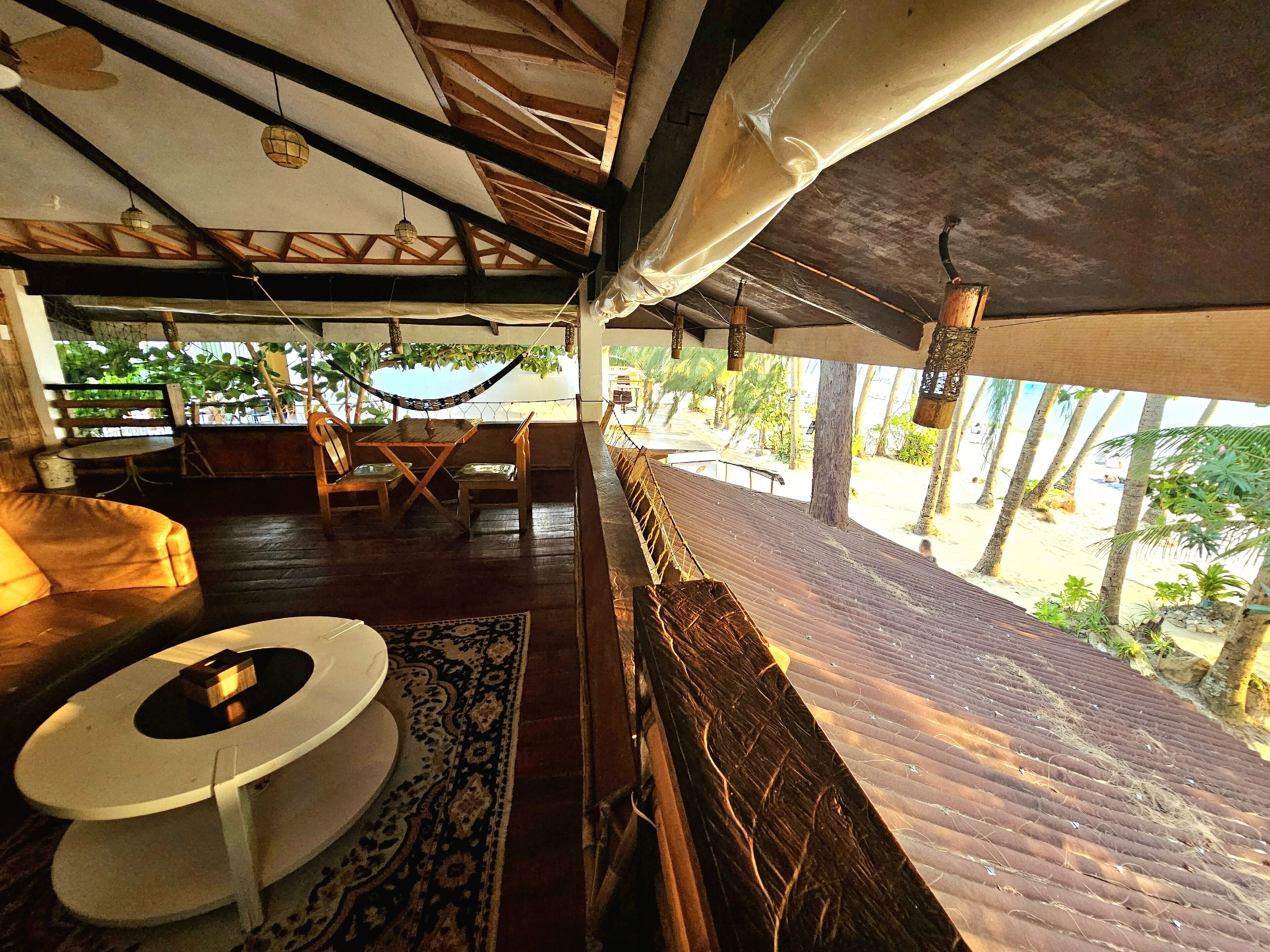 Silla hamaca, mesa de madera y zona de estar con cojines, con vista a una playa y palmeras.