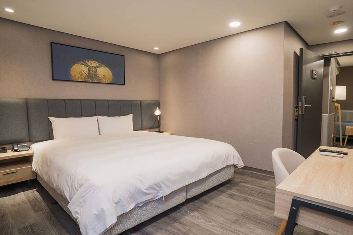 Une chambre d’hôtel avec un grand lit, des tables de chevet, un bureau avec une chaise, un tableau au mur et un miroir dans un coin.