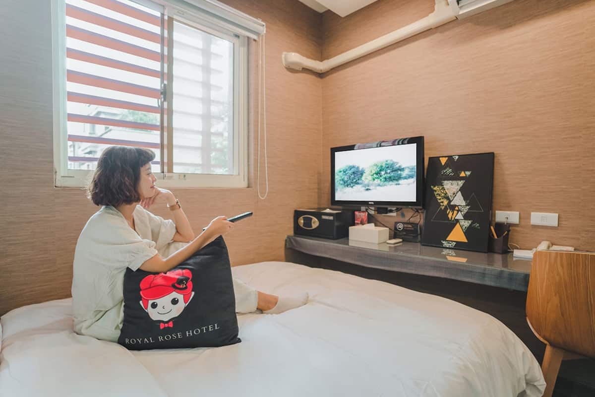 Une personne est assise sur un lit dans une chambre d’hôtel. On aperçoit une fenêtre, un bureau avec un ordinateur et un oreiller orné d’un motif Hello Kitty.