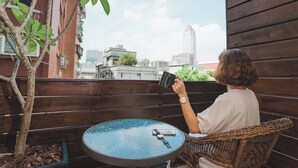 Una persona sentada frente a una mesa redonda, con un teléfono inteligente y una taza, contemplando un paisaje urbano con edificios y un árbol.