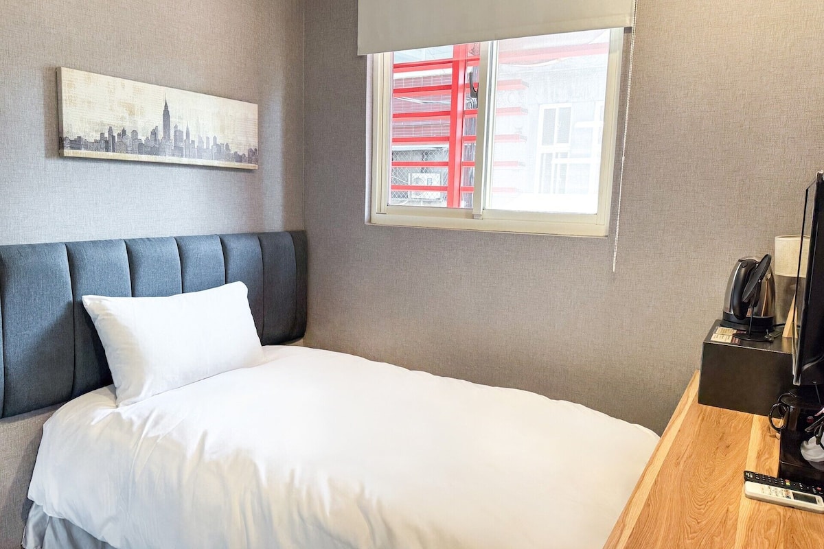 Une chambre d’hôtel avec un lit, une tête de lit, une fenêtre avec un garde-corps rouge, une machine à café et une télécommande posée sur une table de chevet en bois.