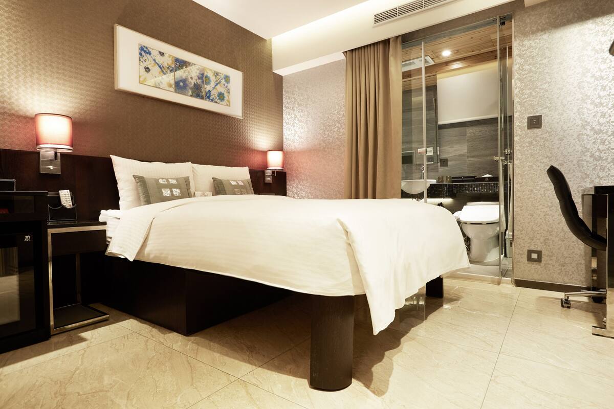 Een moderne hotelkamer met een groot bed, bedlampen, een nachtkastje en een badkamer zichtbaar door een glazen deur.