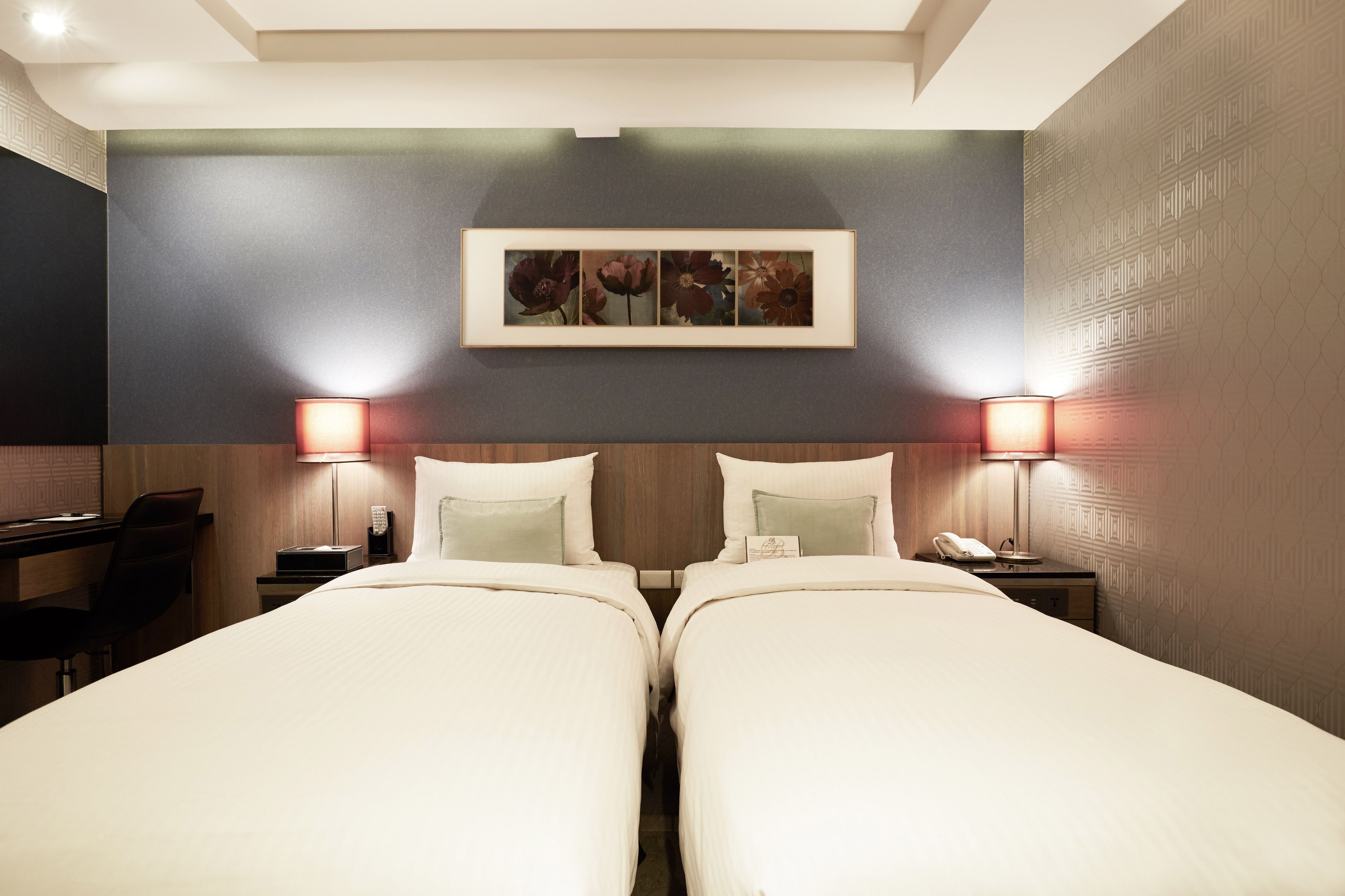 Une chambre d’hôtel avec deux lits, un bureau, une chaise et une œuvre d’art encadrée au-dessus des lits.