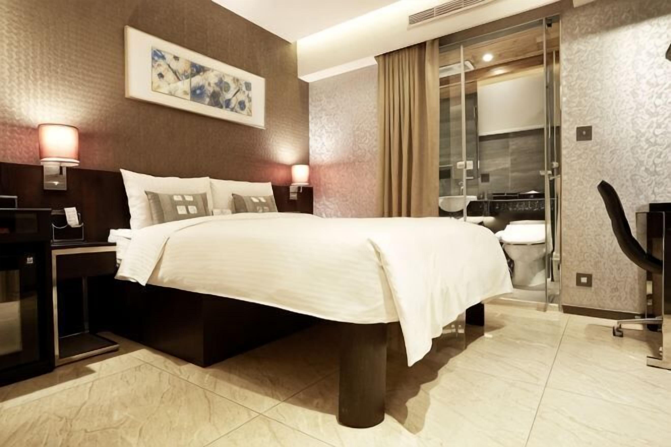 A hotel room with a bed, bedside tables, a desk, a chair, a TV, and a bathroom with a shower and toilet.