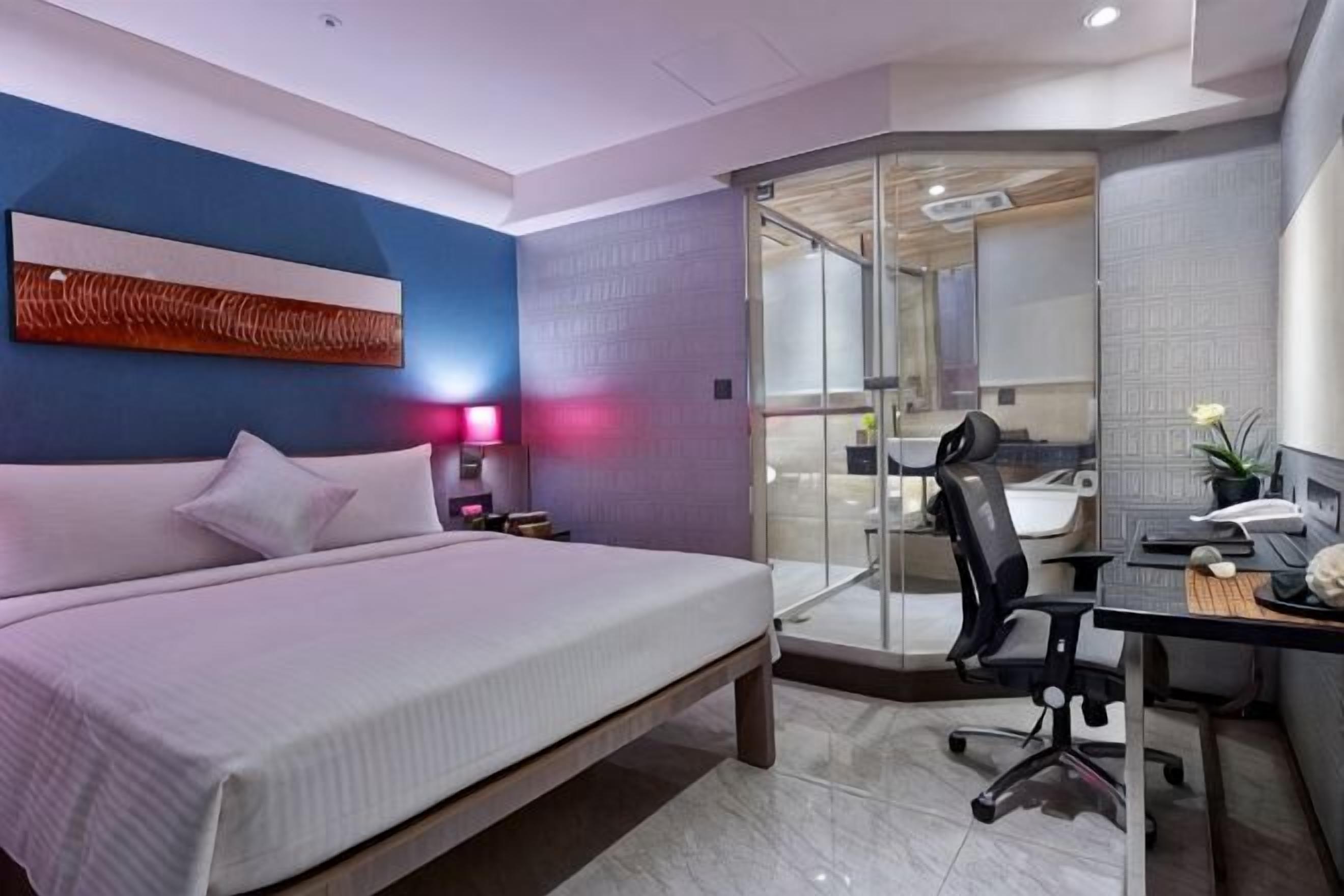 A modern hotel room with a bed, desk, chair, and a glass partition separating the bedroom from the bathroom.