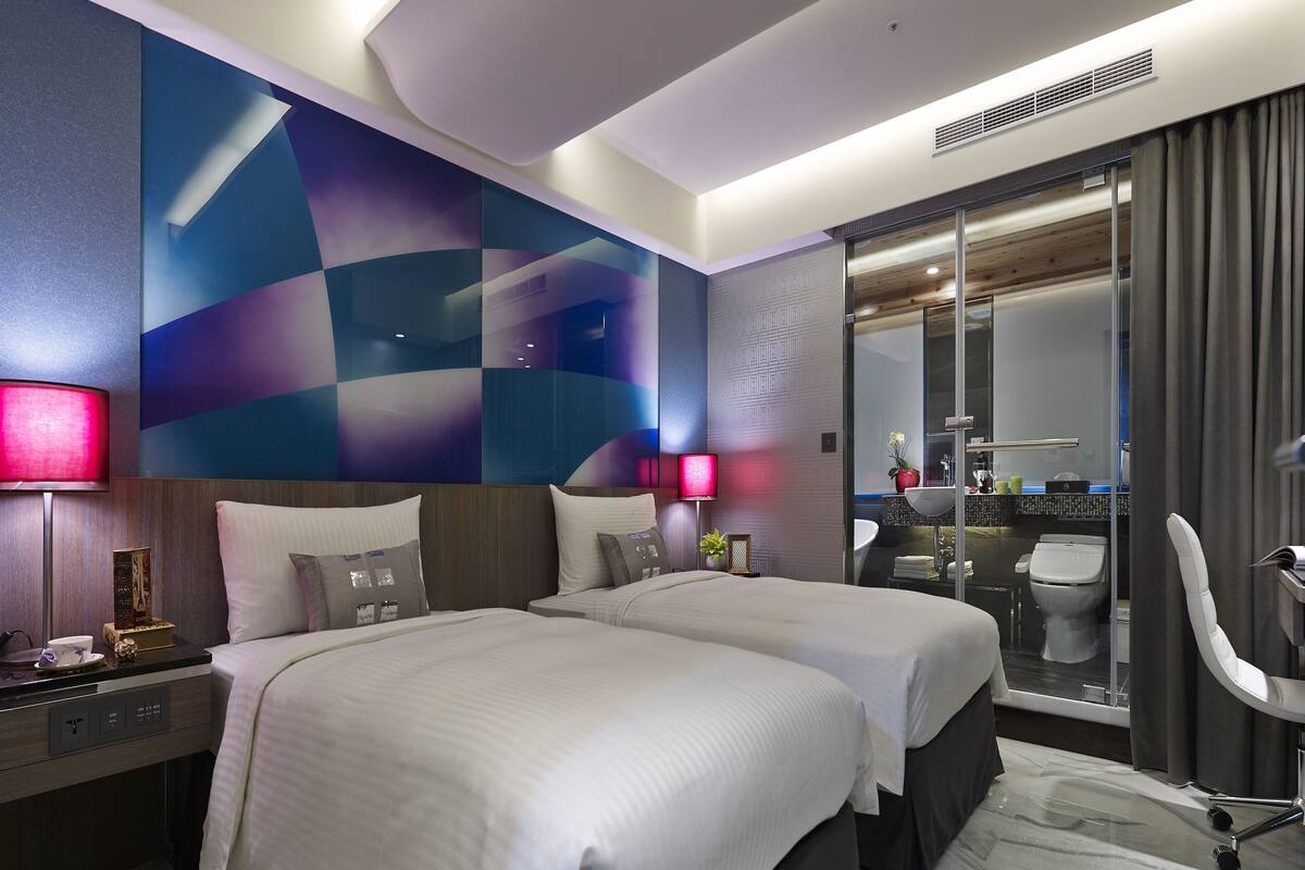 Een hotelkamer met twee bedden, een nachtkastje, een lamp, een televisie en een zichtbare badkamer door een glazen deur.