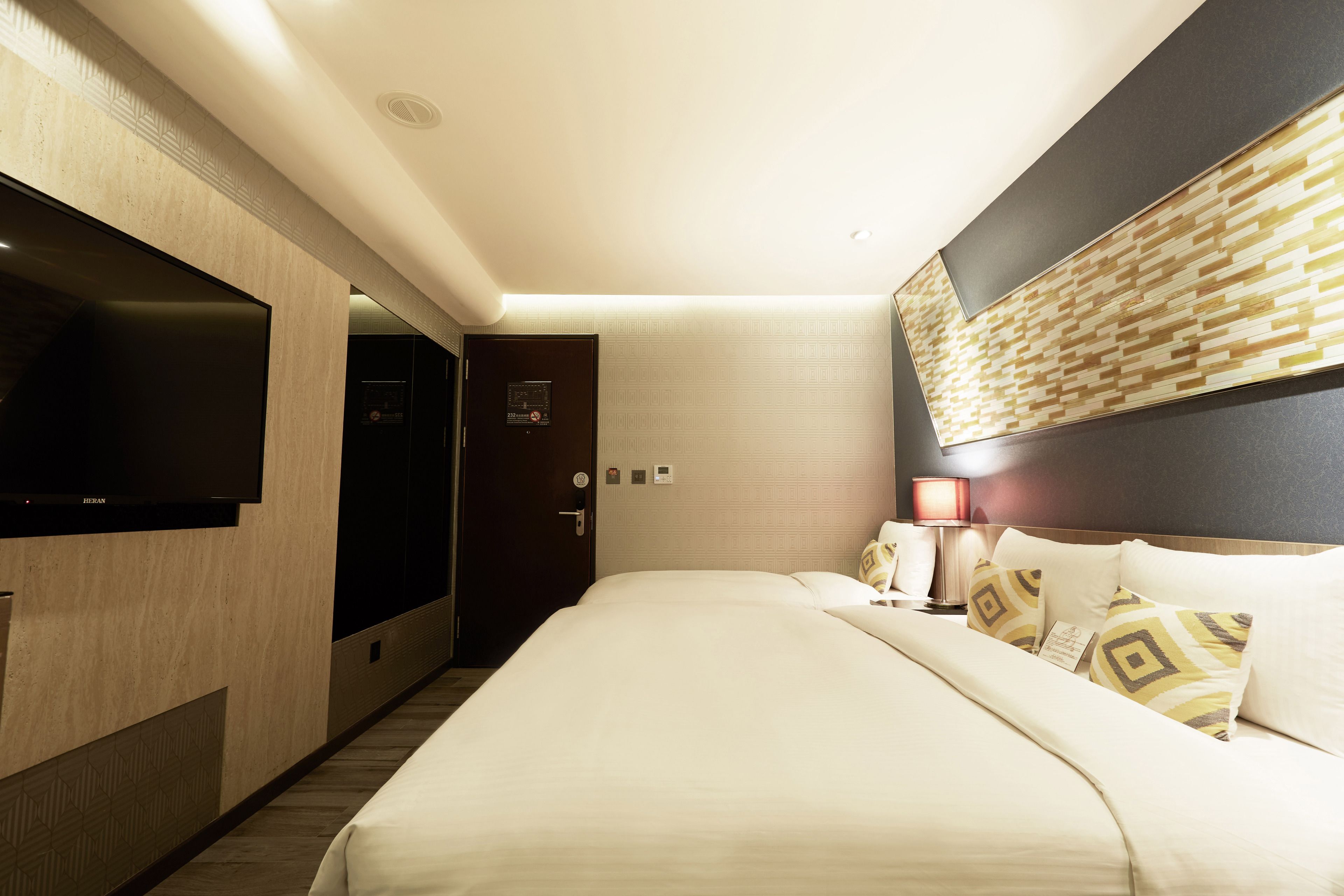 A hotel room with a large bed, a flat-screen TV mounted on the wall, a bedside table with a lamp, and a door leading to a bathroom.