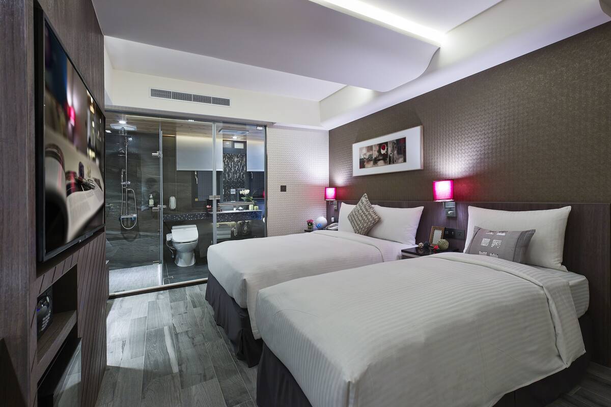 Een hotelkamer met twee bedden, een televisie, een badkamer met douche en een klein tafeltje met lamp.