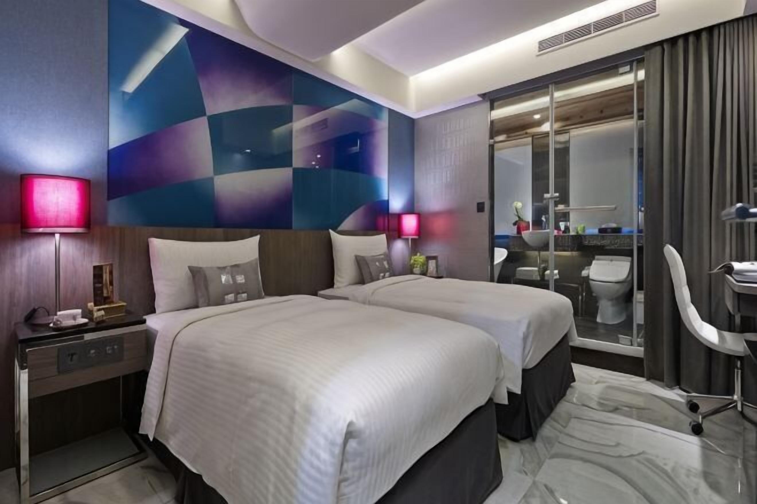 A hotel room with two beds, a desk, a chair, and a bathroom visible through a glass door.