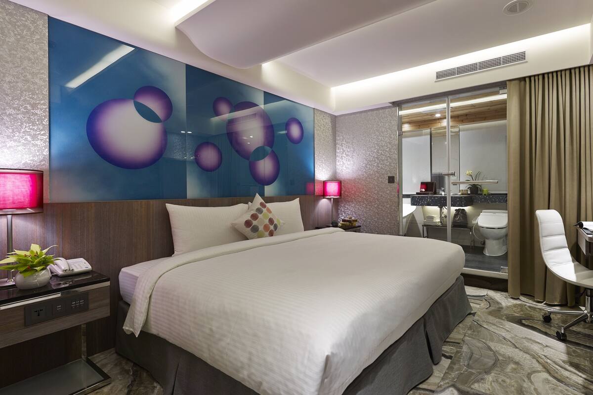 Een moderne hotelkamer met een groot bed, een bureau met een lamp, een spiegel en een zichtbare badkamer achter een glazen deur.