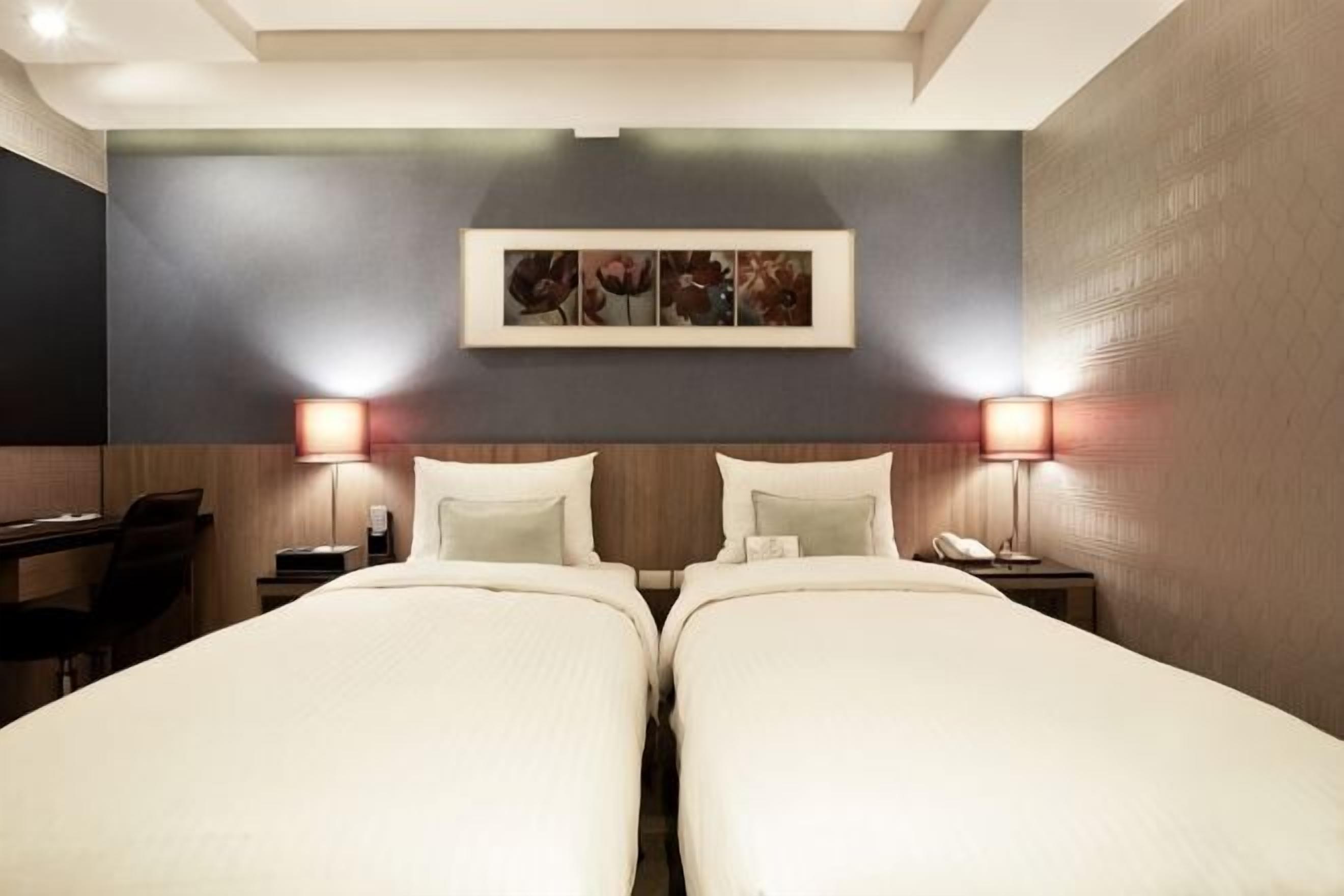 A hotel room with two beds, a desk, a chair, a phone, and a wall-mounted picture.