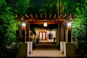 A wooden pergola entrance with a tiled pathway leading to a building, surrounded by lush greenery.