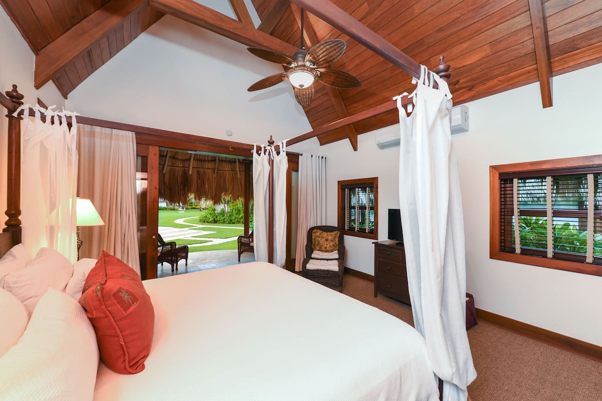 A bedroom with a large bed, a ceiling fan, a view of a garden, and a window with blinds.