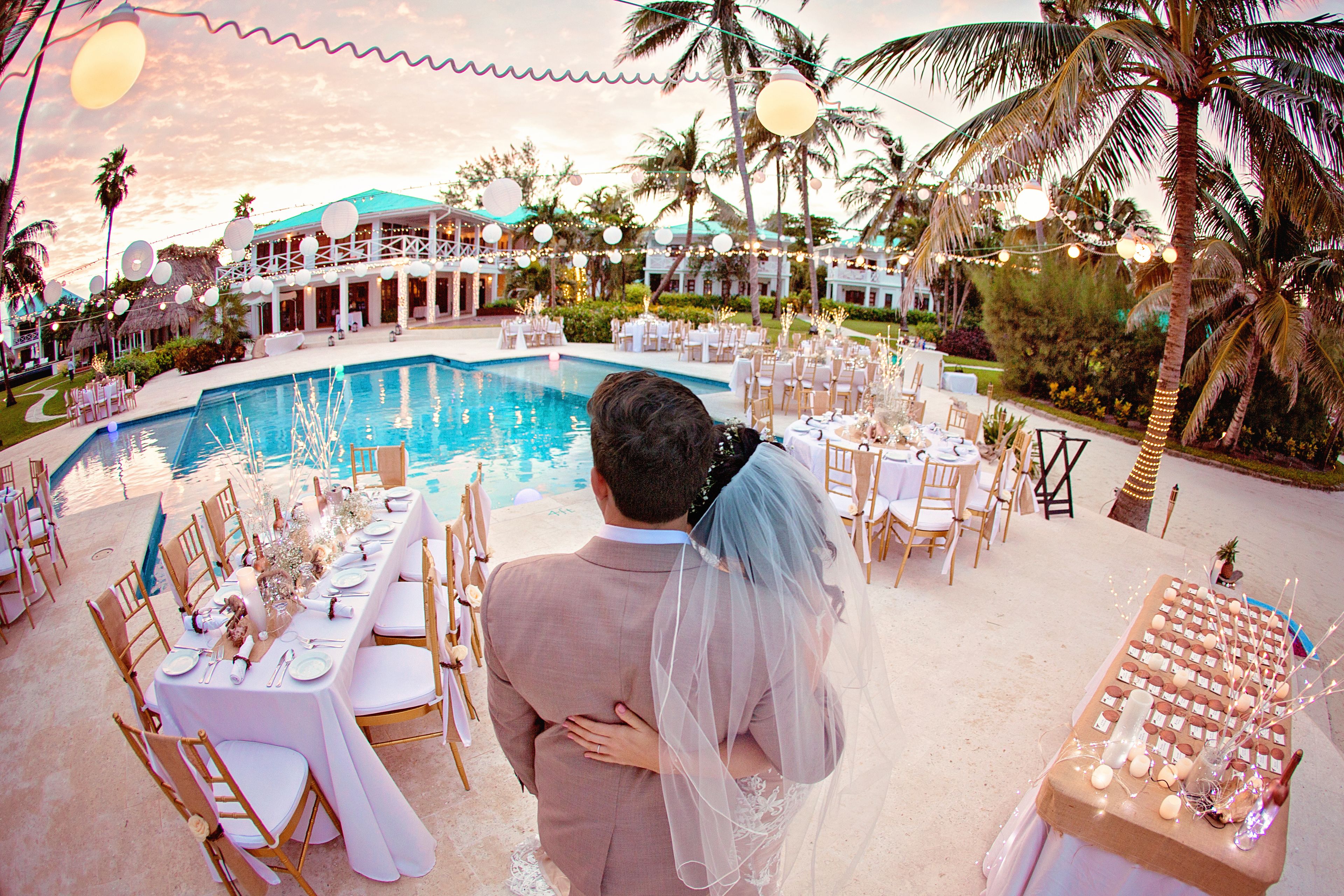 A wedding reception with a pool, tables set for guests, and palm trees in the background.