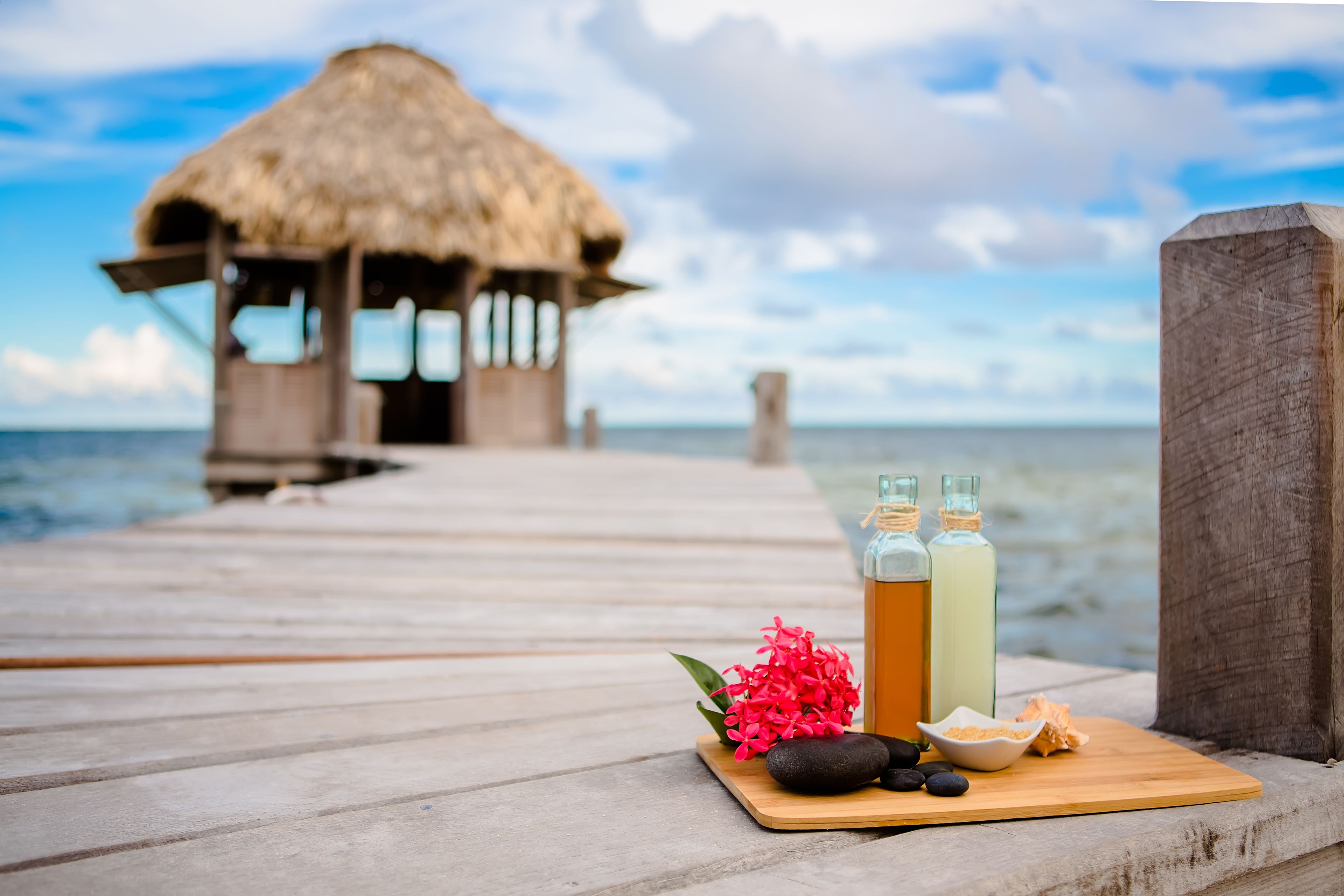 A wooden pier leading to a thatched-roof structure with a wooden table holding a bottle, a bowl, and a flower.
