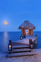 Una estructura de madera con techo de paja sobre un muelle, con vista al mar, y una luna llena al fondo.