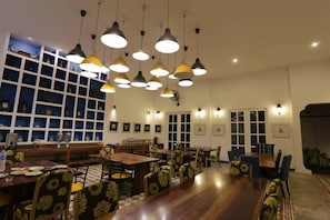 Un comedor con varias mesas y sillas, iluminación colgante y una pared de estanterías con diversos objetos.