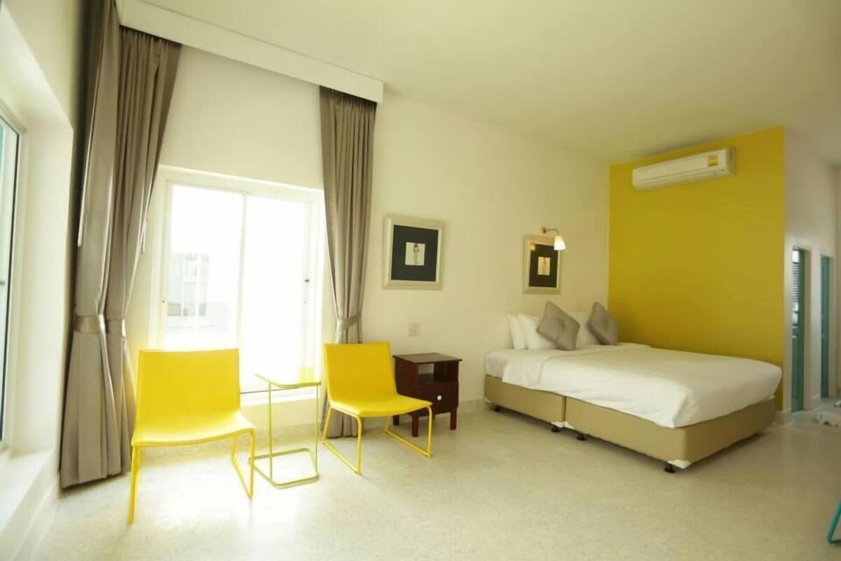 A hotel room with a bed, two yellow chairs, a small table, a window with curtains, and an air conditioning unit.