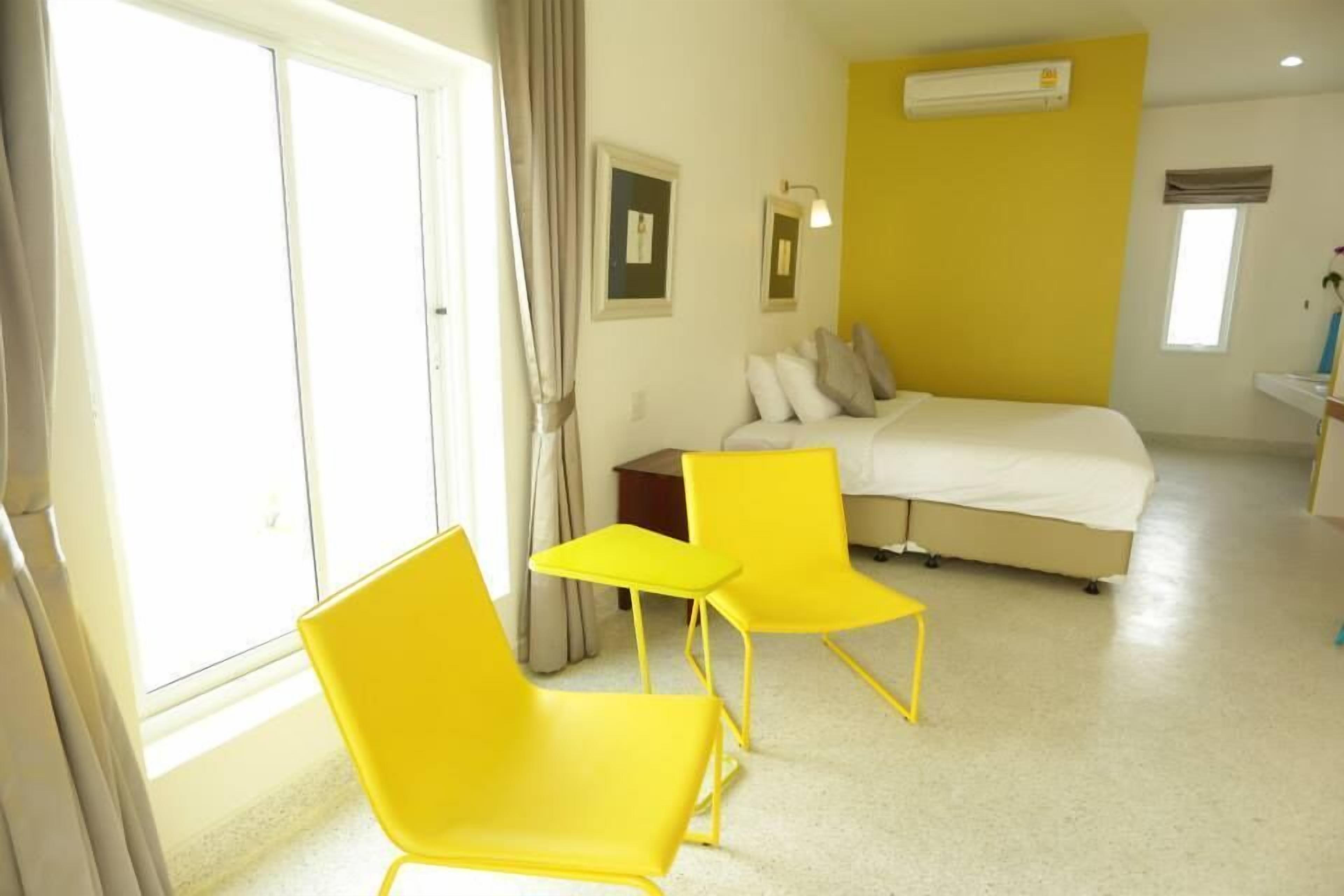 Una habitación de hotel con una cama, dos sillas amarillas, una mesita, una ventana con cortinas y un equipo de aire acondicionado en la pared.