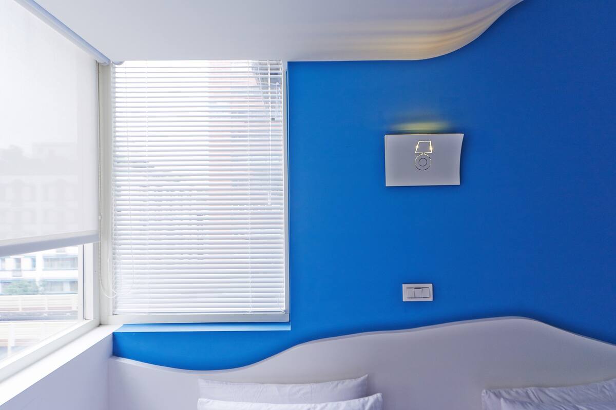 A room with a blue wall, a white bed, and a window with blinds.