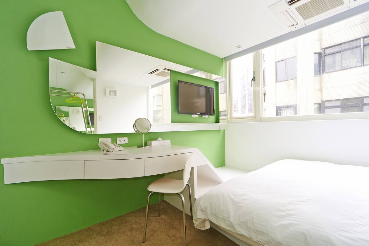 A bedroom with a green wall, a white bed, a desk with a chair, a TV, and a window.