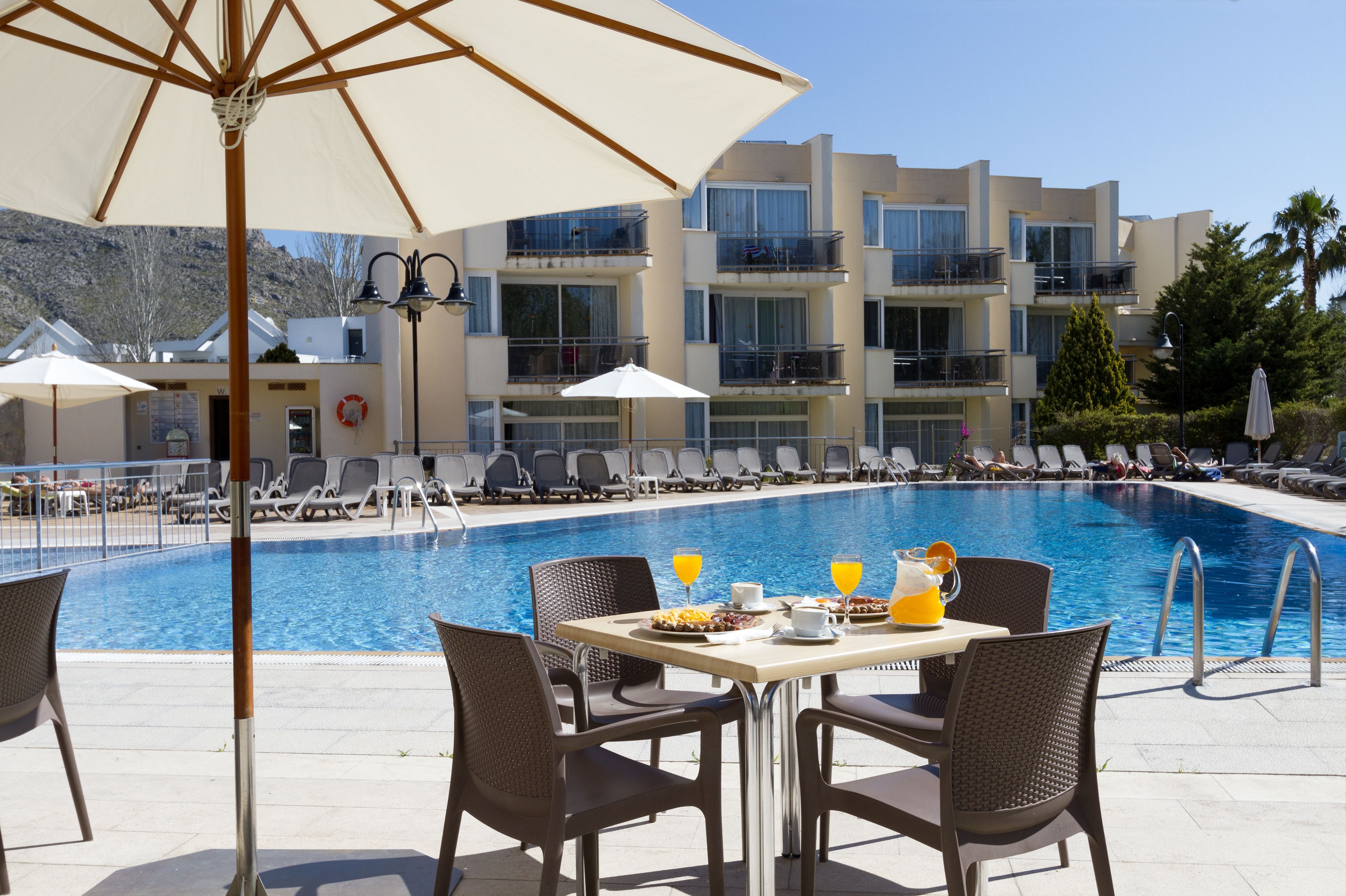 A poolside area with lounge chairs, a table with breakfast items, and a building with balconies in the background.