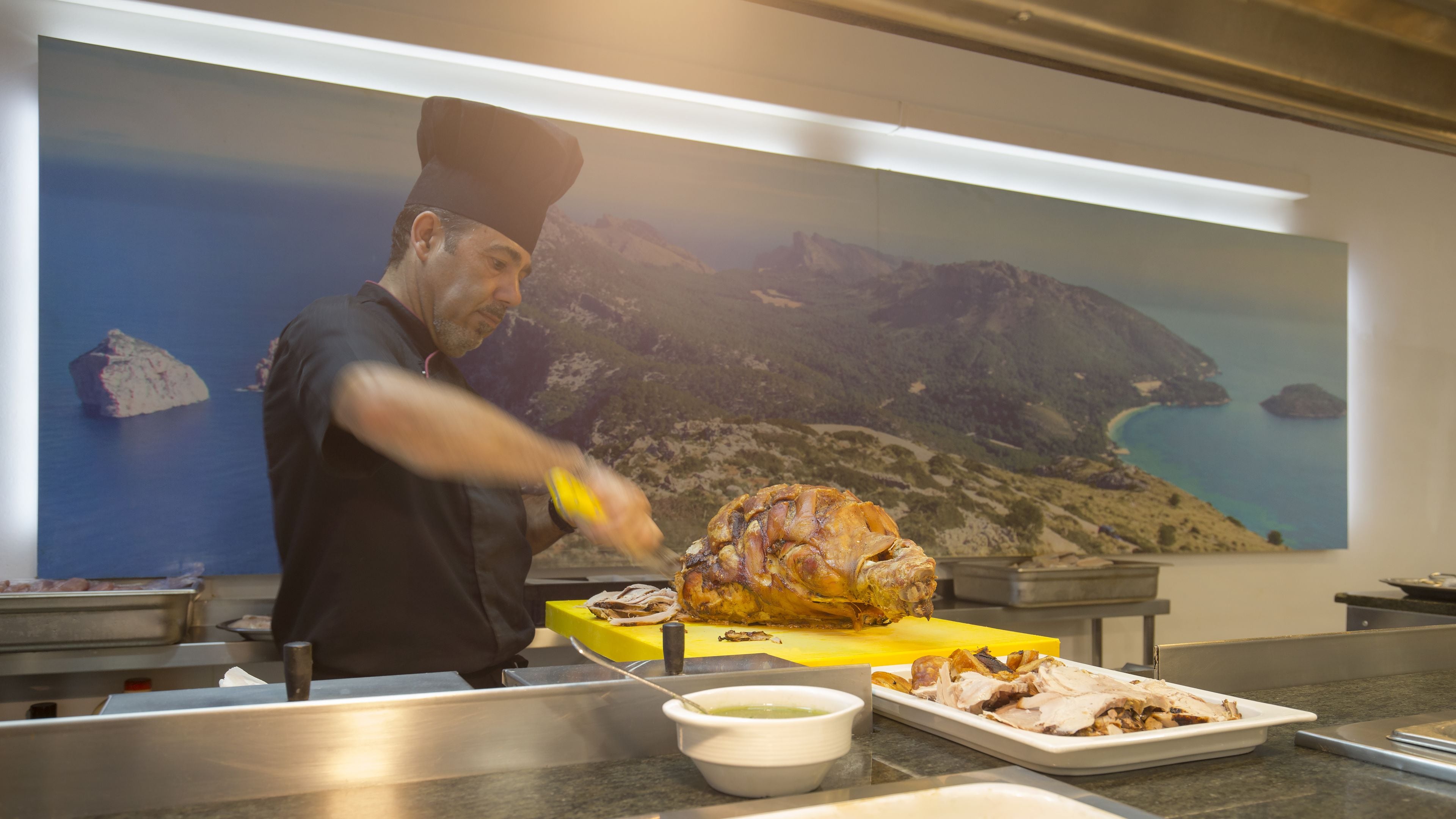 A chef carving a large roasted turkey on a yellow cutting board in a kitchen with a scenic coastal mural in the background.