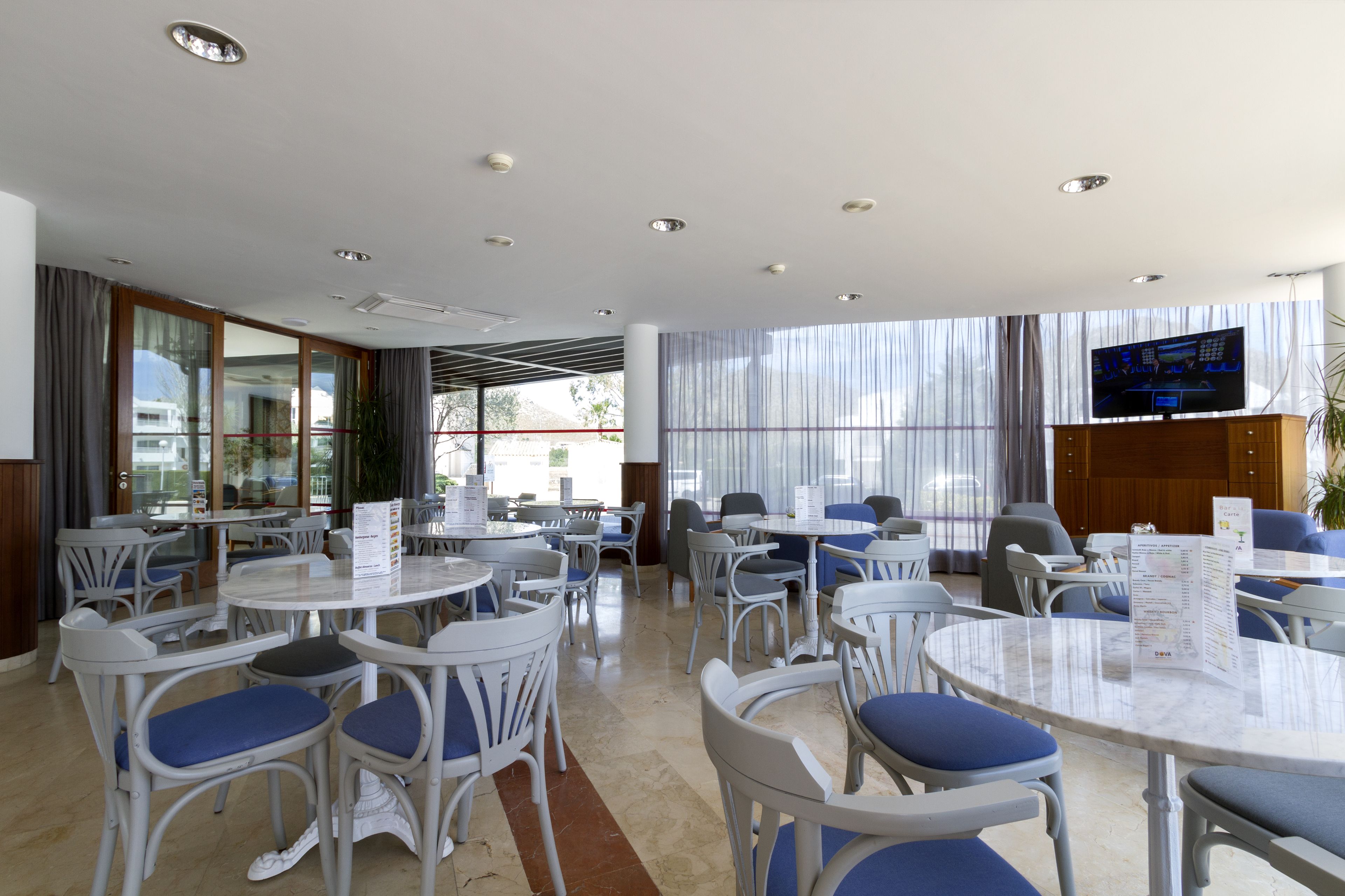 A hotel dining area with round tables and chairs, a TV mounted on the wall, and large windows with curtains.