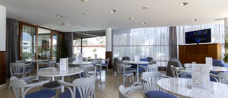Un comedor de hotel con mesas y sillas redondas, un televisor en la pared y ventanas amplias con cortinas.