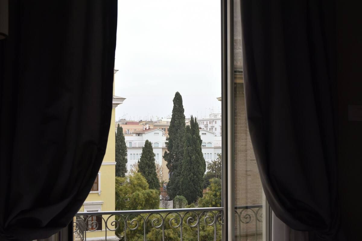 A view from a balcony with curtains, overlooking a row of buildings and tall trees.