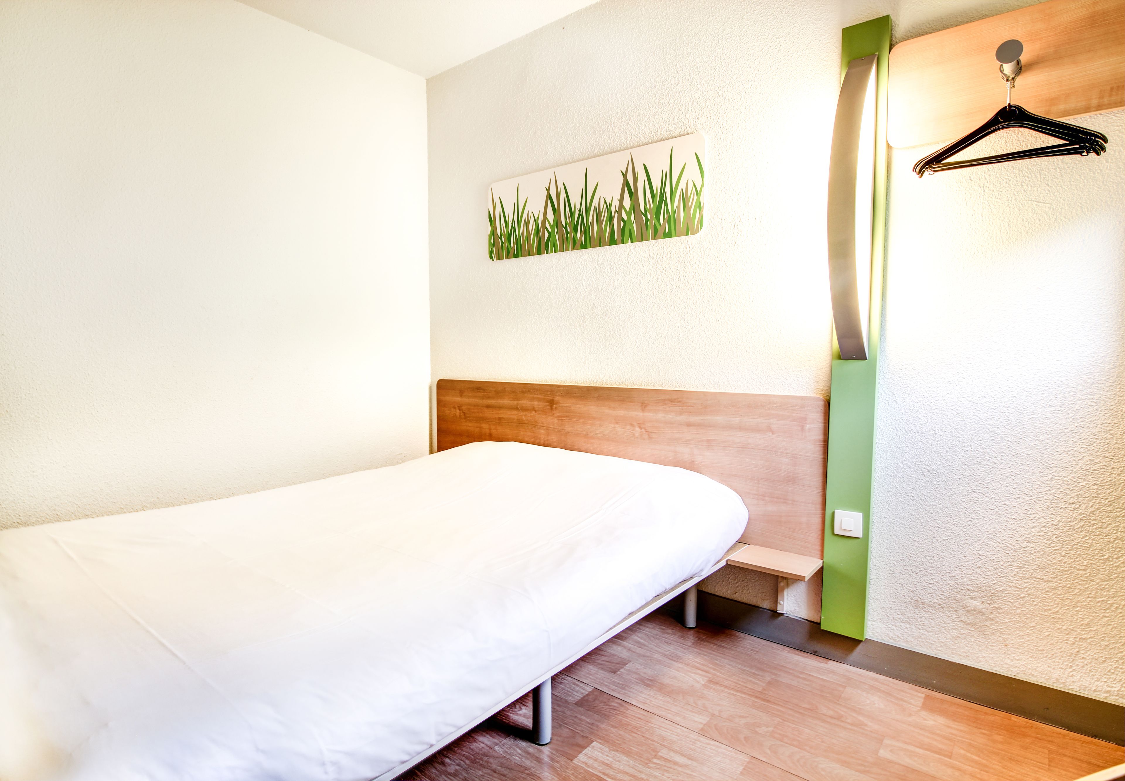 Una cama individual en una habitación con cabecera de madera, luz adosada a la pared y una pared de acento verde.
