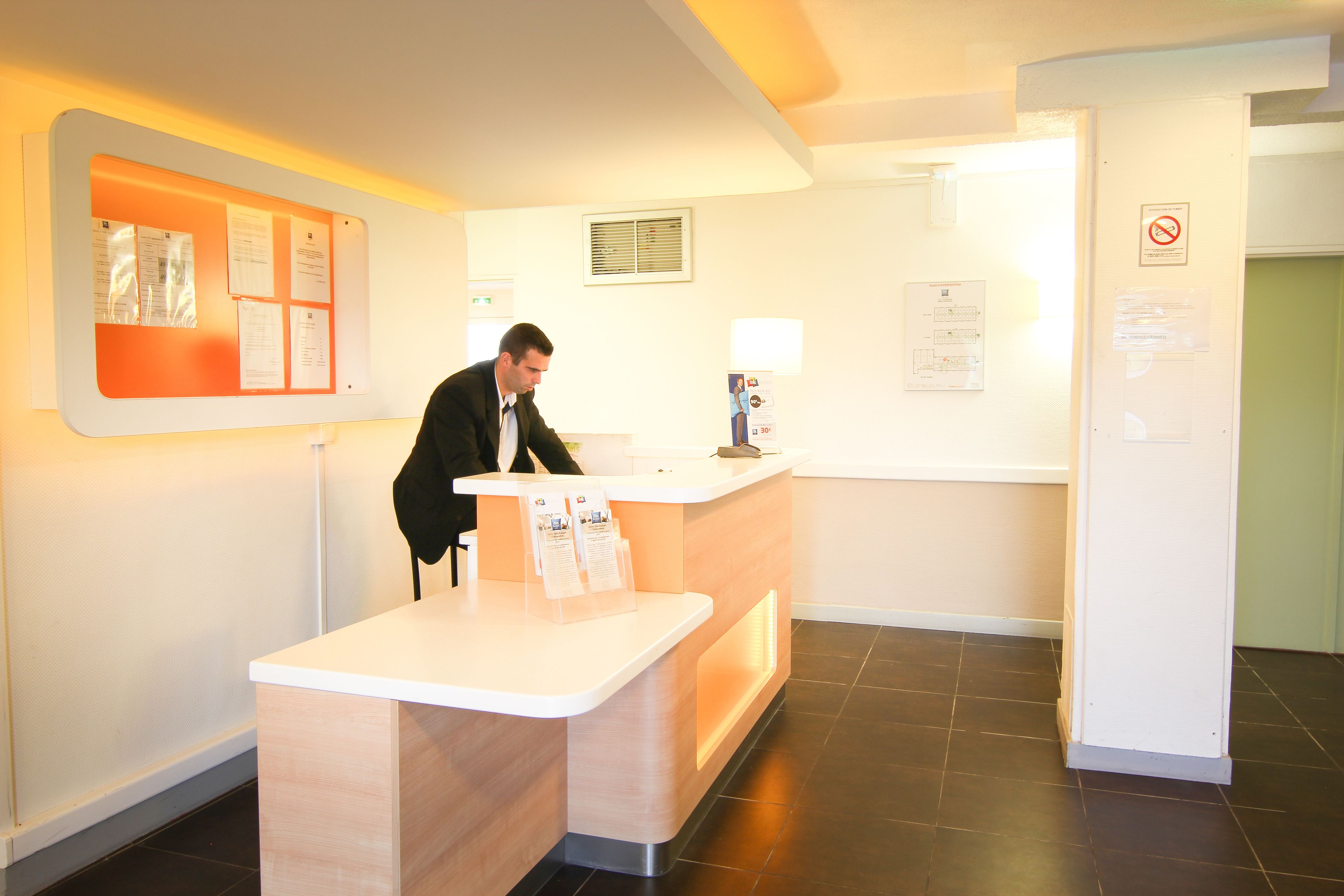 A reception desk with a man standing behind it, a bulletin board with papers, and a no-smoking sign on the wall.