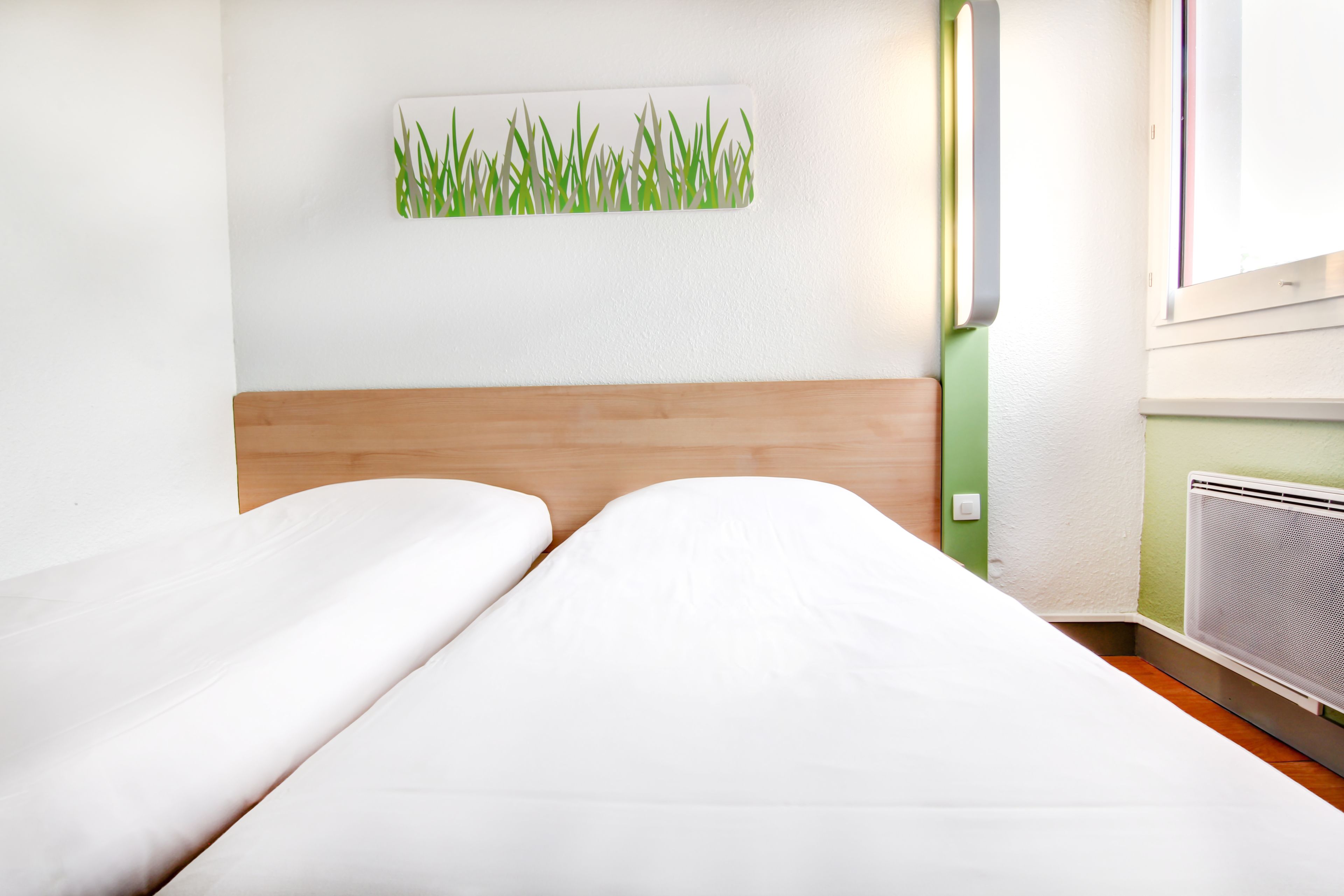 Une chambre d’hôtel avec deux lits, une tête de lit en bois, une œuvre d’art représentant de l’herbe accrochée au mur et un radiateur.