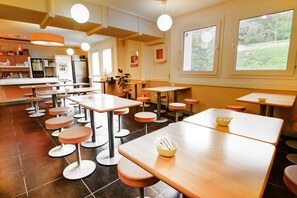 A restaurant with multiple tables and stools, a kitchen area, and large windows with a view of greenery. - ibis budget Chateaudun (Chateaudun)