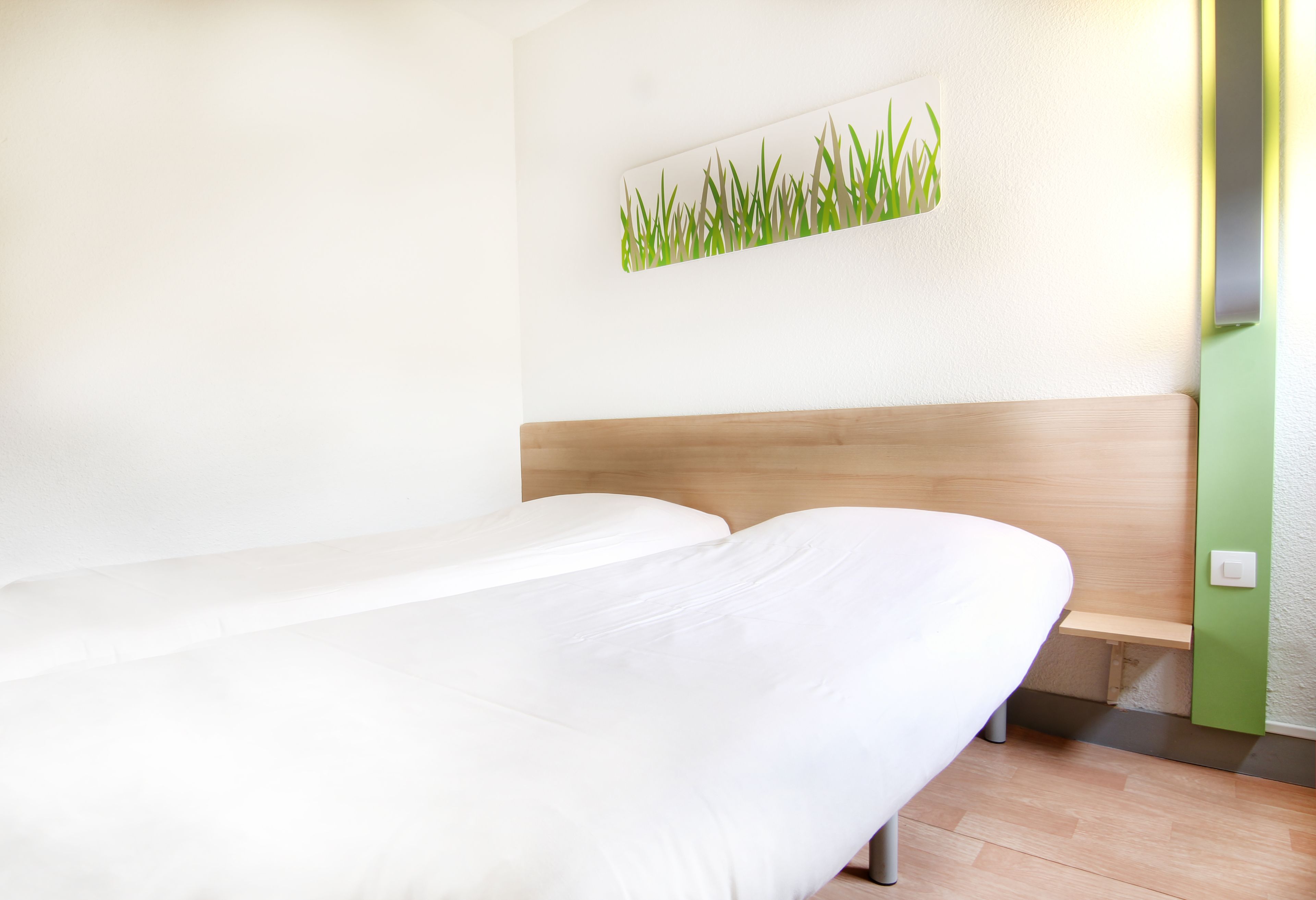 A hotel room with a single bed, a wooden headboard, a small bench, and a wall-mounted artwork of grass.