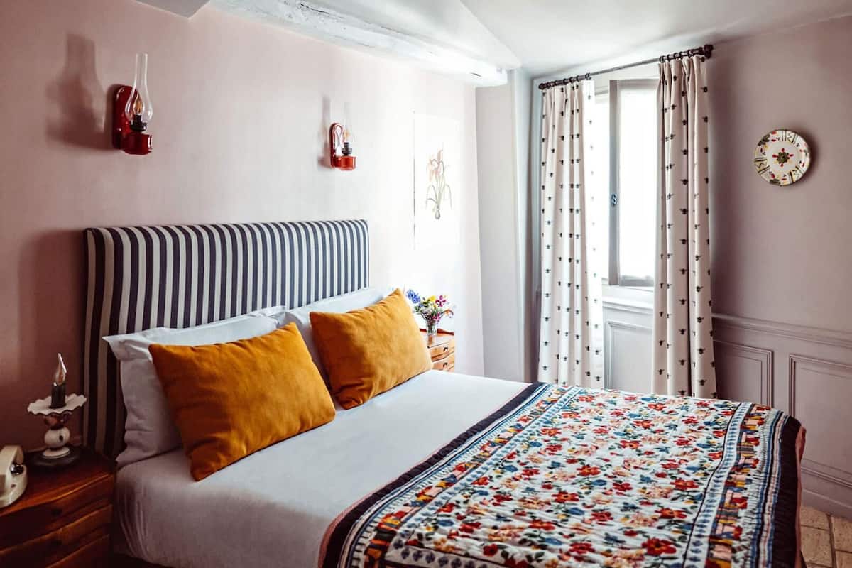 Une chambre à coucher avec une tête de lit à rayures, deux oreillers, une couvre-lit à motifs floraux, une table de chevet avec une lampe et une fenêtre avec des rideaux.